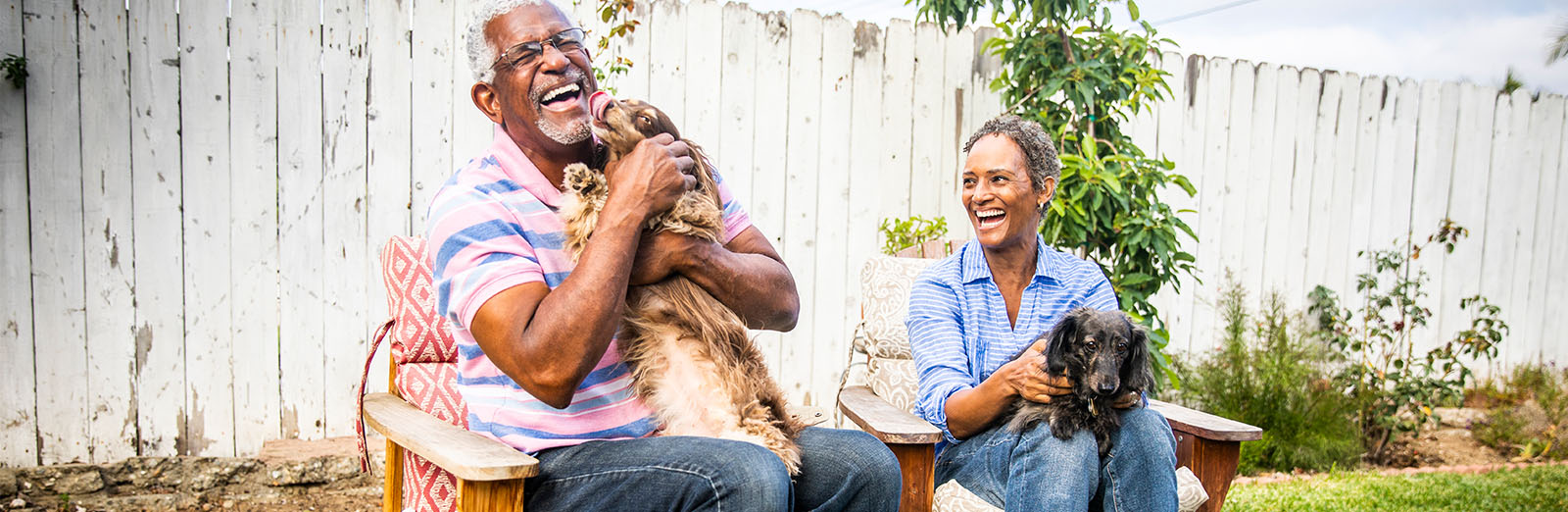 A couple sitting outside and holding dogs. The dog is licking the face of the man.