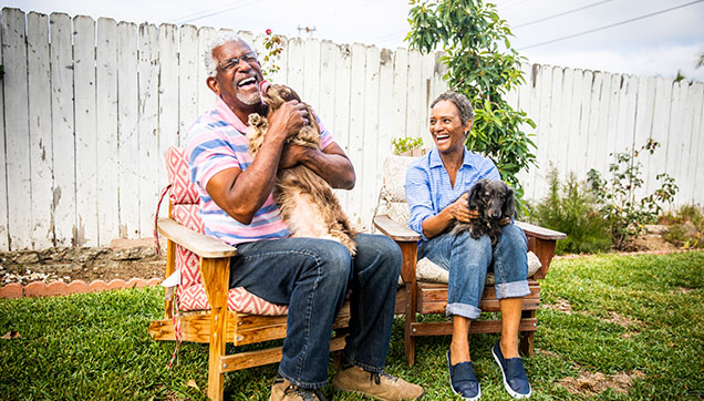 A couple sitting outside and holding dogs. The dog is licking the face of the man.