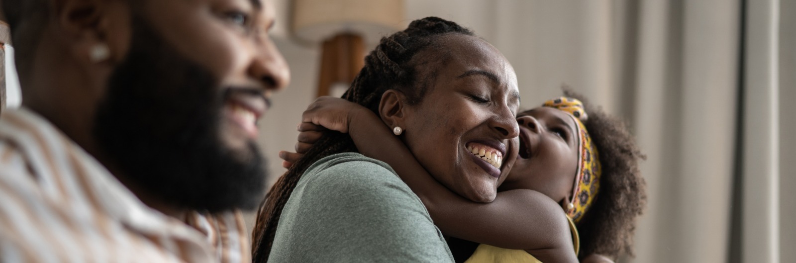 A family sitting in a living room and a daughter has her arms wrapped around her mother's neck