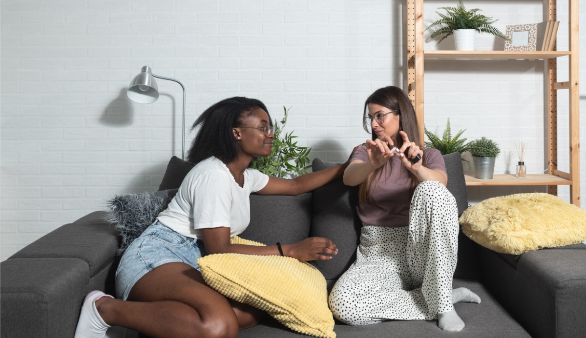 Two women sitting on a couch and one is breaking a cigarette in half.