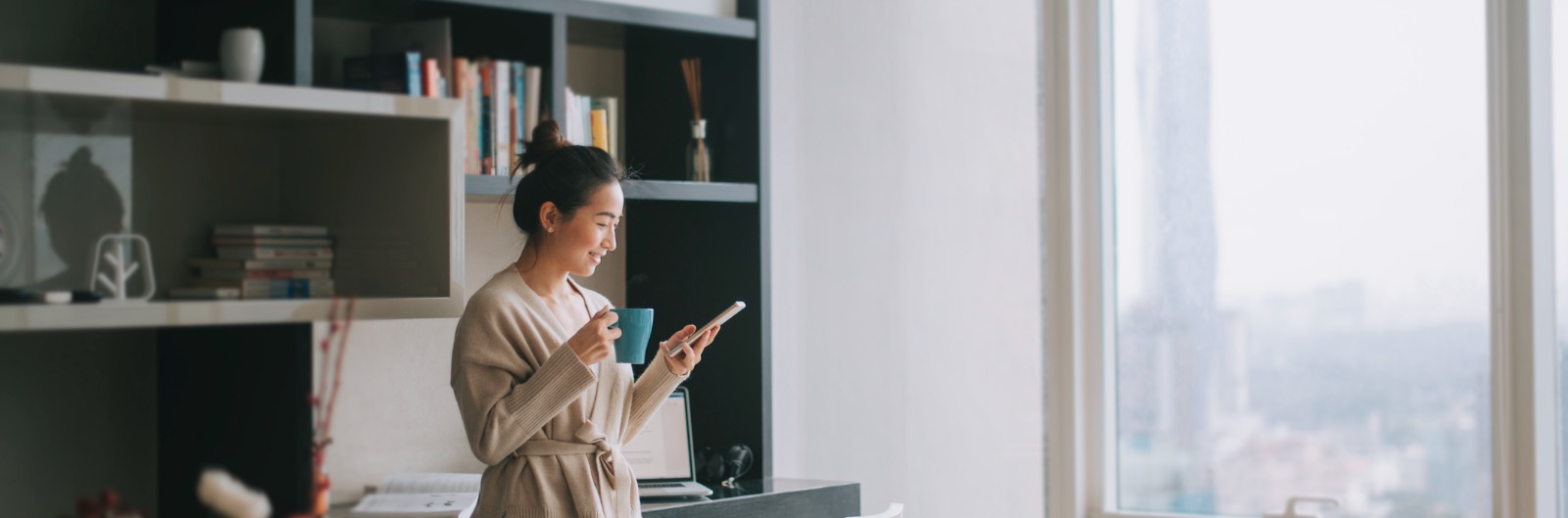 A woman standing in a sky rise condo and looking at her phone while holding a cup